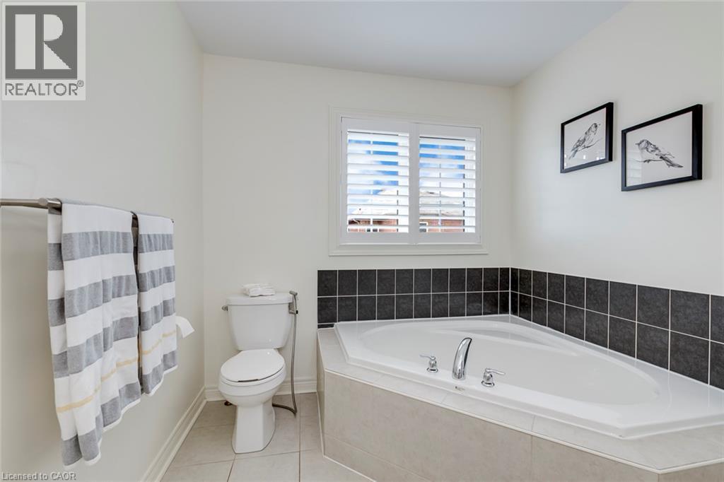 Corner soaking tub with polished chrome fixtures, white window shutters, dark tile wainscoting, white toilet, and neutral floor tiling - 598 Bessborough Drive, Milton, ON - Indoor Photo Showing Bathroom