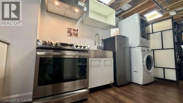 Stainless steel range with ventilation hood, stainless steel sink, under-sink cabinetry, stacked washer and dryer, and wood-finish flooring - 598 Bessborough Drive, Milton, ON - Indoor Photo Showing Laundry Room