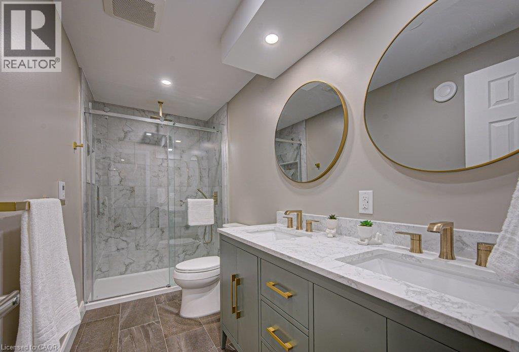 Dual vanity with brass hardware and round mirrors, a glass-enclosed shower with a rainfall showerhead, full-height marble-patterned tile, and wood-finish flooring - 15 Matheson Crescent, Innerkip, ON - Indoor Photo Showing Bathroom