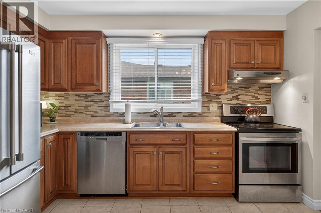 Kitchen featuring warm wood cabinetry, stainless steel appliances, a mosaic tile backsplash, a double basin sink, and neutral tile flooring - 56 Brookstream Court, Hamilton, ON - Indoor Photo Showing Kitchen With Stainless Steel Kitchen With Double Sink