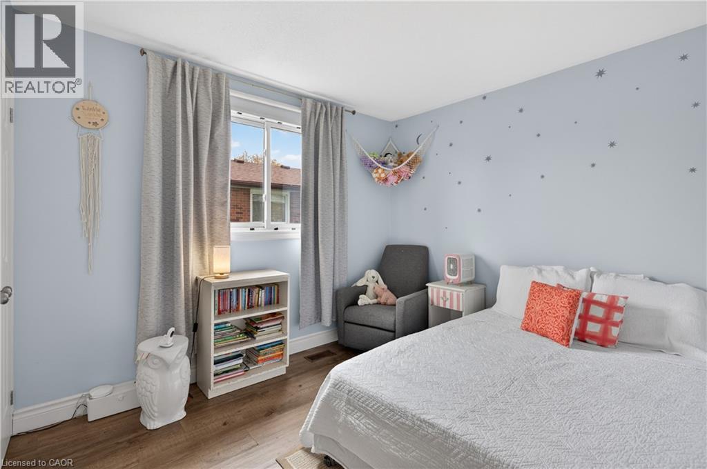 Bedroom featuring light blue walls, wood-finish flooring, a double window with light-filtering curtains, white baseboards, and a built-in wall heater - 56 Brookstream Court, Hamilton, ON - Indoor Photo Showing Bedroom