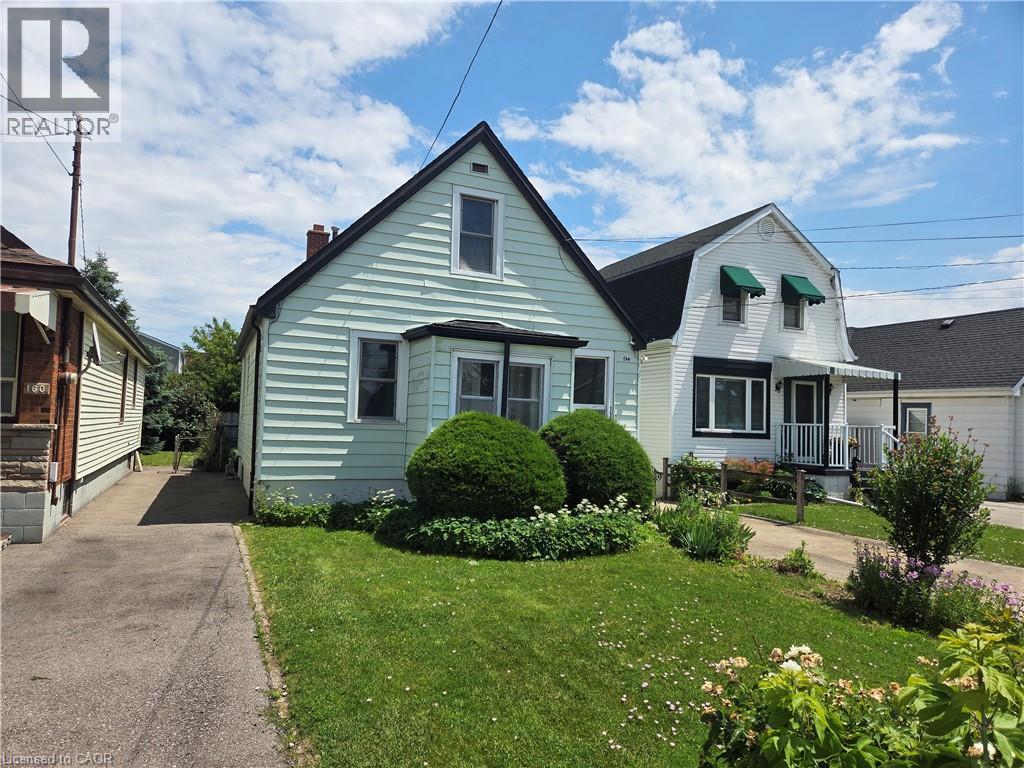 Light-colored siding exterior featuring a contrasting dark roof, bay window, and a manicured lawn with established plantings - 156 Adeline Avenue, Hamilton, ON