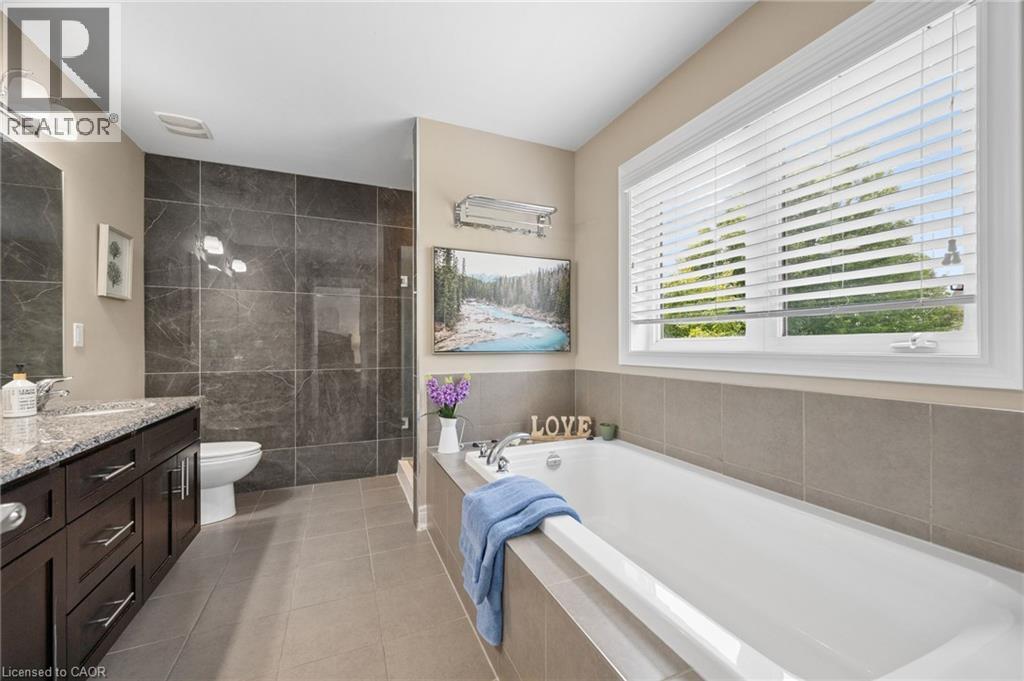 Spacious bathroom featuring a large soaking tub, separate glass-enclosed shower with dark stone-finish tiling, a vanity with ample storage, a stone-finish countertop, and a window with blinds - 145 Redtail Street, Kitchener, ON - Indoor Photo Showing Bathroom