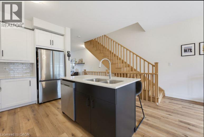 Kitchen featuring a contemporary black island with an undermount double sink, stainless steel appliances, white cabinetry, and wood-finish flooring - 16 Raspberry Lane, Hamilton, ON - Indoor Photo Showing Kitchen With Double Sink