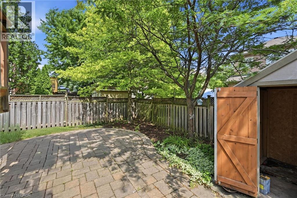 Paved patio area featuring interlocking pavers, a privacy fence, mature trees, and a utility shed with a wooden door - 5628 Stella Lane, Burlington, ON - Outdoor