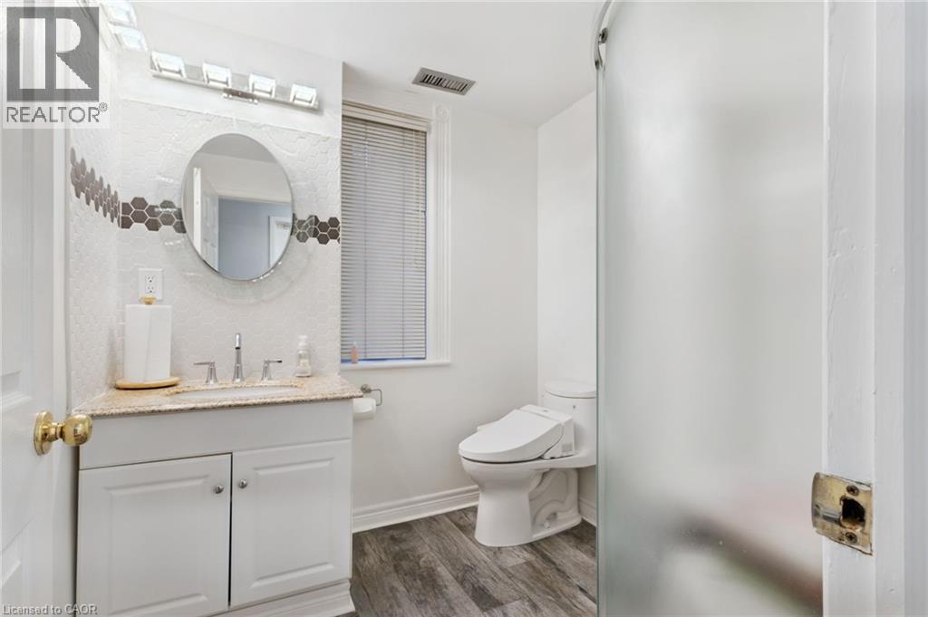 Bathroom featuring a Shower, white vanity with a granite-style countertop, a circular mirror, and a wall-mounted light fixture - 189 Hughson Street, Hamilton, ON