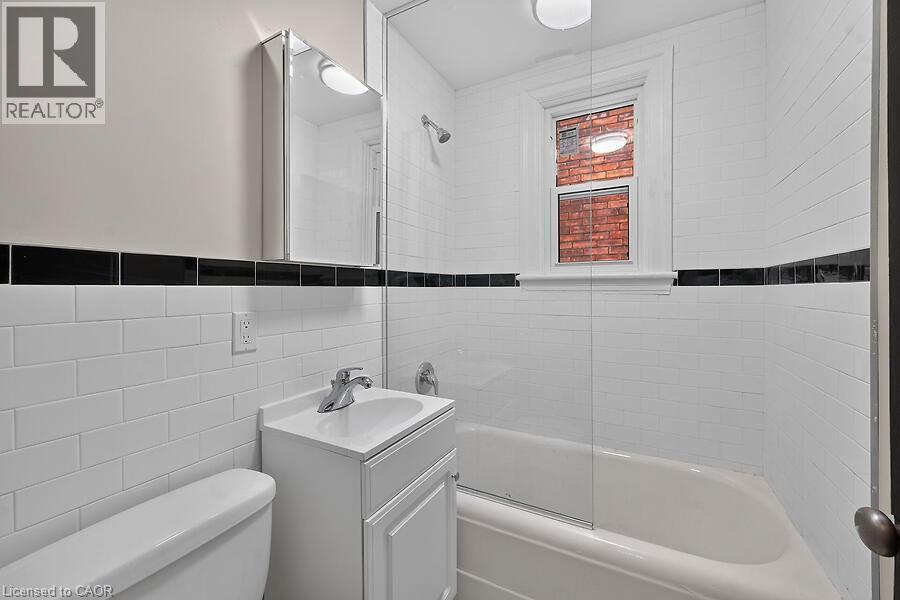 Bathroom featuring white subway tile walls with a black accent band, a white vanity with an integrated sink, a mirrored medicine cabinet, a bathtub with a clear glass shower enclosure, and a window pr - 103 Mary Street, Hamilton, ON - Indoor Photo Showing Bathroom