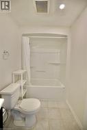 Full bathroom featuring a white tub and shower combination with integrated shelving, a white toilet, and light-toned tile flooring -
