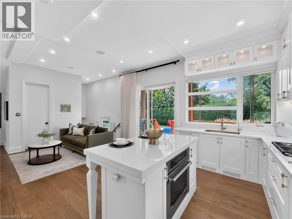 Open-concept kitchen featuring an integrated oven, white cabinetry with gold-tone hardware, a large window overlooking green foliage, and wood-finish flooring - 5178 Hunter Drive, Burlington, ON - Indoor Photo Showing Kitchen With Upgraded Kitchen