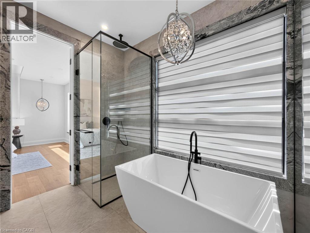 Freestanding soaking tub with a matte black faucet, glass-enclosed shower, large format tile flooring, stone-clad walls, and a decorative crystal chandelier - 5178 Hunter Drive, Burlington, ON - Indoor