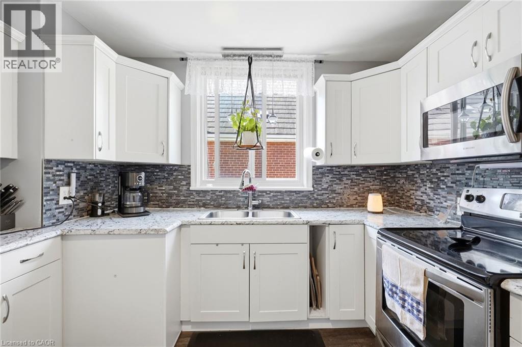 Bright kitchen featuring white cabinetry, a mosaic tile backsplash, granite-finish countertops, stainless steel appliances, and a double basin sink beneath a window - 127 Taylor Avenue, Hamilton, ON - Indoor Photo Showing Kitchen With Double Sink