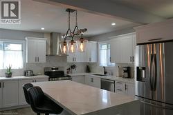 Contemporary kitchen featuring white shaker cabinetry, stainless steel appliances, a large center island with a light-toned countertop, and a designer pendant light fixture - 