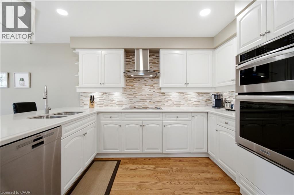 Bright kitchen featuring white cabinetry, light-toned countertops, stainless steel appliances, wood-finish flooring, and a textured tile backsplash - 1090 Crofton Way, Burlington, ON - Indoor Photo Showing Kitchen With Double Sink With Upgraded Kitchen