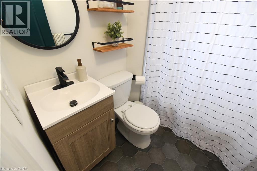 Bathroom featuring hexagonal floor tiles, a wood-finish vanity with a white countertop and integrated sink, a matte black faucet, and a round wall-mounted mirror - 106 Belview Avenue, Hamilton, ON - Indoor Photo Showing Bathroom