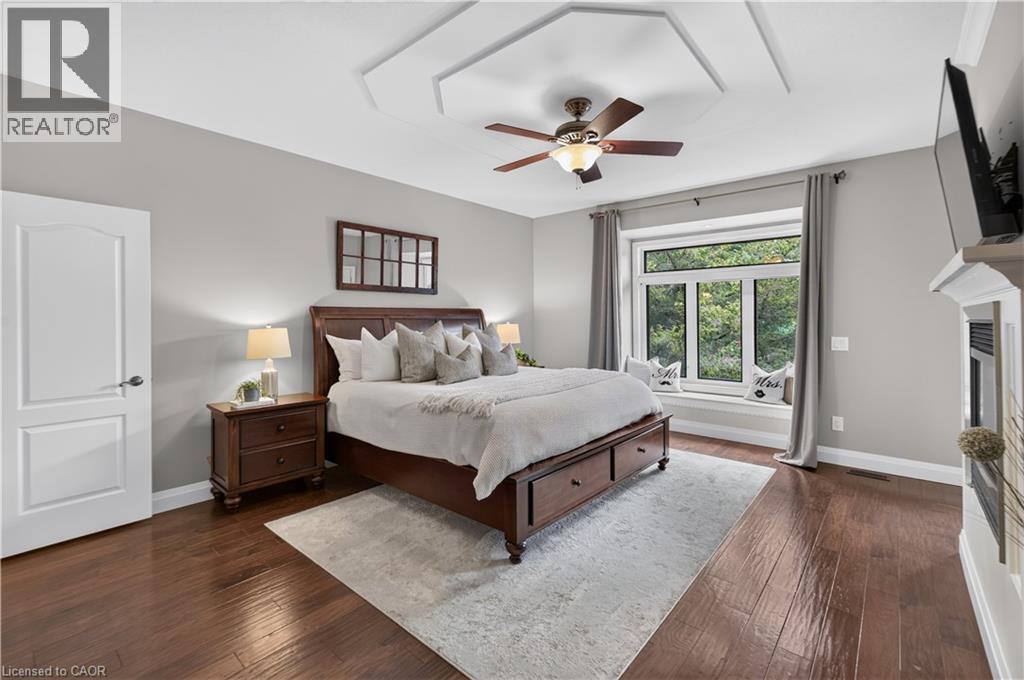Hardwood flooring throughout, featuring a tray ceiling with a ceiling fan, a window seat beneath a large window, a built-in fireplace, and a two-panel interior door - 2050 15 Side Road, Milton, ON - Indoor Photo Showing Bedroom
