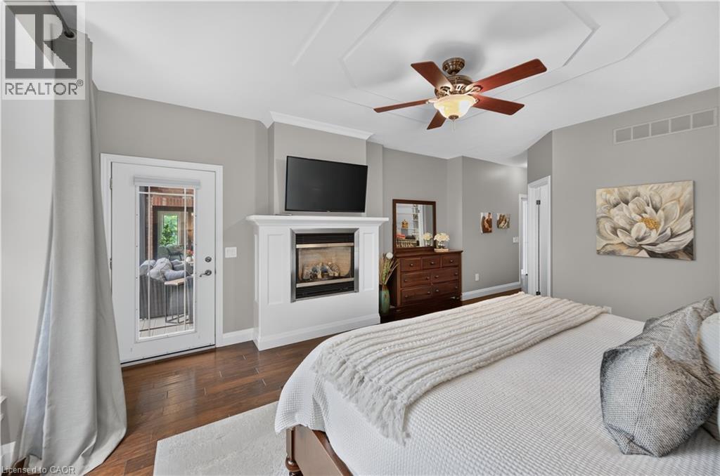 Spacious room featuring wood-finish flooring, a tray ceiling with a ceiling fan, a fireplace with a white mantel, and a glass-paned door - 2050 15 Side Road, Milton, ON - Indoor Photo Showing Bedroom With Fireplace