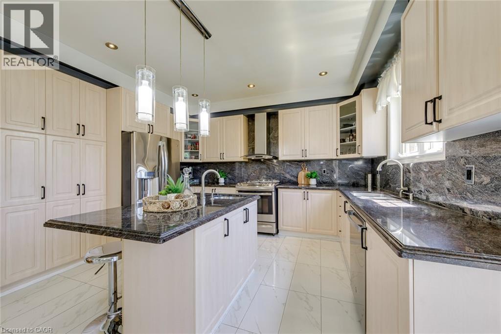 Contemporary kitchen featuring cream-toned cabinetry, dark stone countertops, stainless steel appliances, a center island with pendant lighting, and white tile flooring - 2380 Woodcrest Drive, Oakville, ON - Indoor Photo Showing Kitchen With Upgraded Kitchen