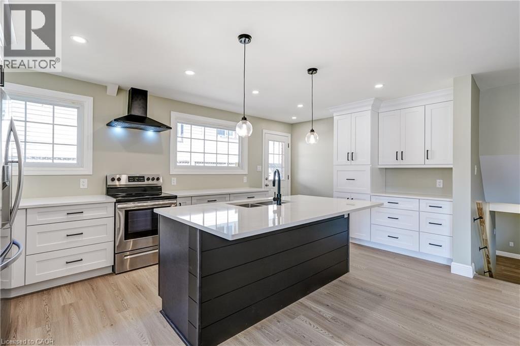 Modern kitchen featuring a dark-toned island with a light-toned countertop, pendant lighting, stainless steel appliances, white cabinetry with black hardware, and wood-finish flooring - 191 Glenmor Drive, Caledonia, ON - Indoor Photo Showing Kitchen With Upgraded Kitchen