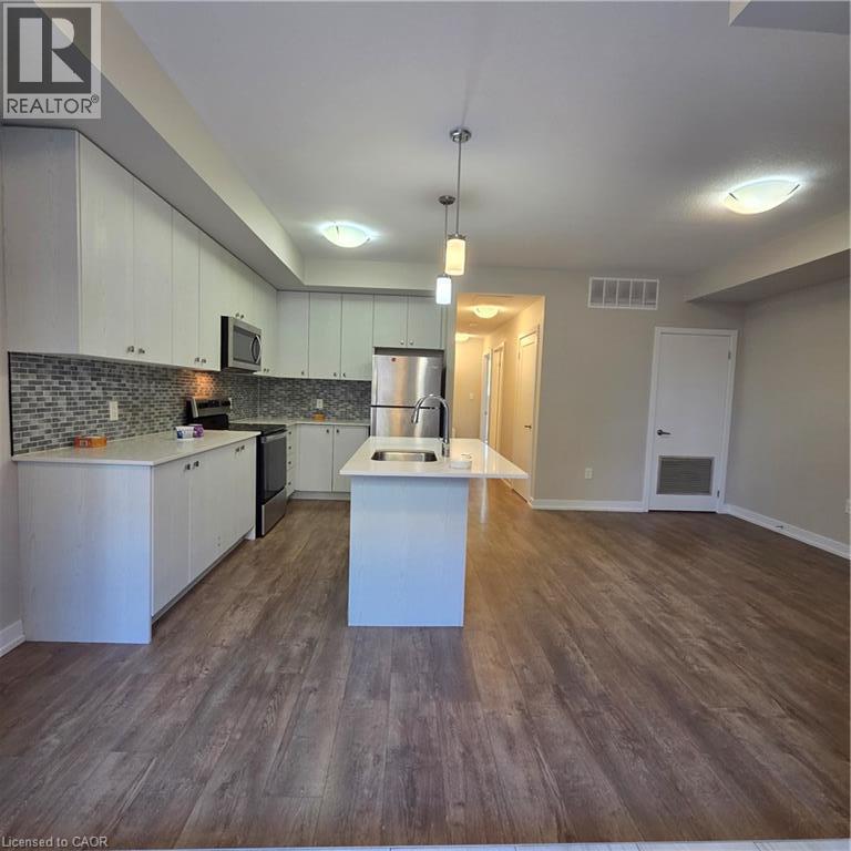 Open-concept kitchen featuring white cabinetry, stainless steel appliances, a mosaic tile backsplash, and an island with an undermount sink - 20 Palace Place Unit# E9, Kitchener, ON - Indoor Photo Showing Kitchen