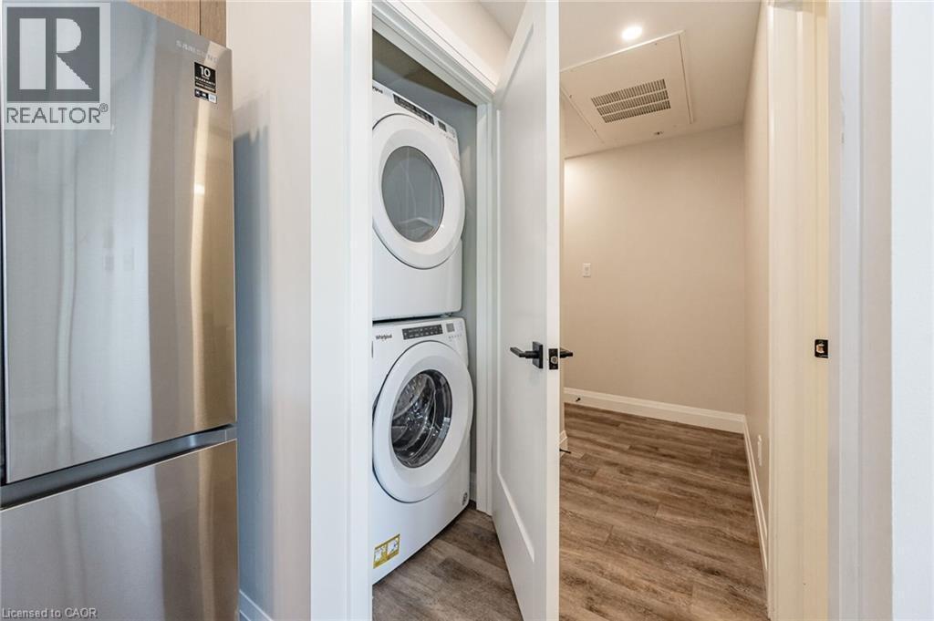 Dedicated laundry closet featuring a stackable washer and dryer, complemented by wood-finish flooring and a contemporary black door handle - 72 Erb Street E Unit# 103, Waterloo, ON - Indoor Photo Showing Laundry Room