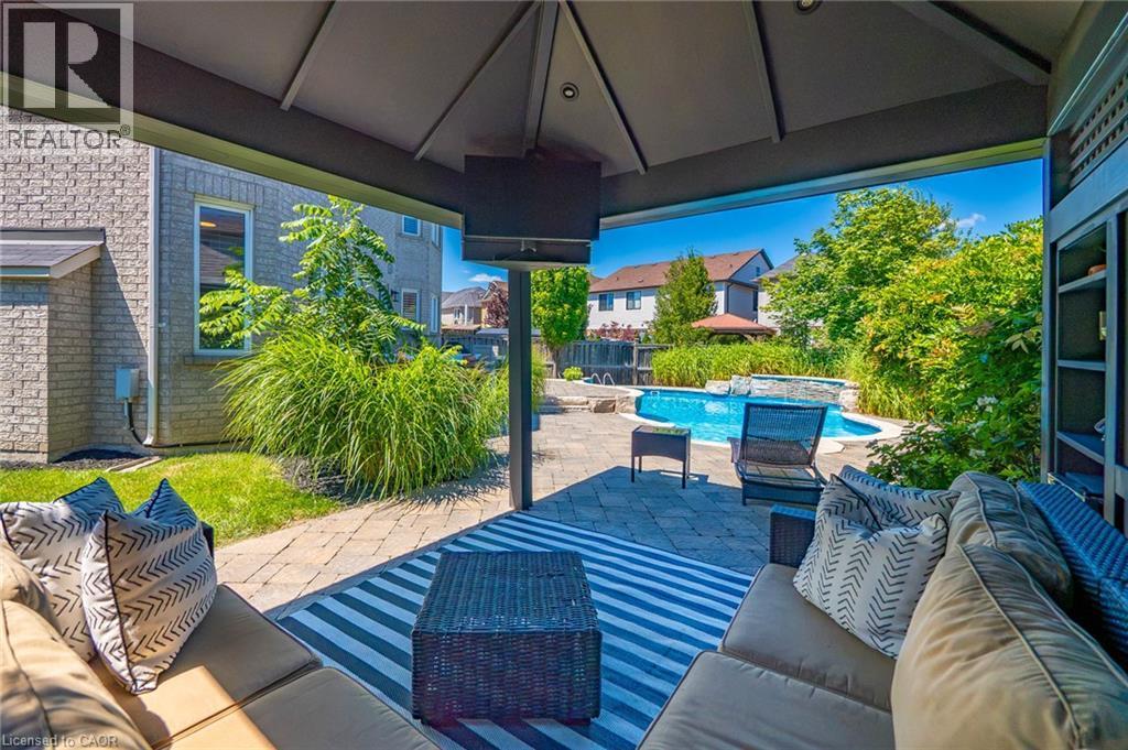 Covered outdoor living space featuring a vaulted ceiling with integrated ceiling fans, striped area rug, built-in seating with cushions, and direct sightlines to the resort-style pool and mature lands - 1533 Morse Place, Milton, ON - Outdoor With In Ground Pool With Deck Patio Veranda