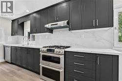 Modern kitchen featuring dark cabinetry, white countertops, a stainless steel range with a double oven, a matching range hood, and a white tile backsplash -
