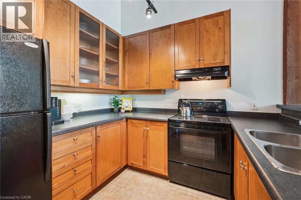 Kitchen featuring warm wood cabinetry, dark countertops, a black electric range, a stainless steel double sink, and a black refrigerator - 26 Ontario Street Unit# 127, Guelph, ON - Indoor Photo Showing Kitchen With Double Sink