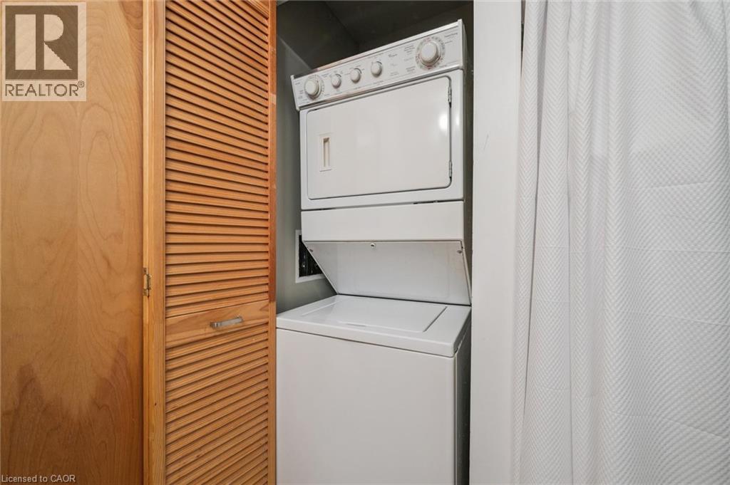White stackable washer and dryer unit, featuring a top-loading washer and front-loading dryer, encased by louvered bi-fold doors with a wood-finish - 26 Ontario Street Unit# 127, Guelph, ON - Indoor Photo Showing Laundry Room