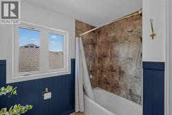 Bathroom featuring a white bathtub with a tiled shower surround, a white framed window, and blue wainscoting -