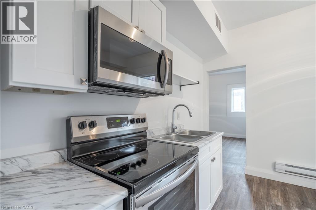 Kitchen featuring white cabinetry, stainless steel microwave, black electric range, dual basin stainless steel sink with a high-arc faucet, and wood-finish flooring - 524 King Street E Unit# 3, Hamilton, ON - Indoor Photo Showing Kitchen With Double Sink