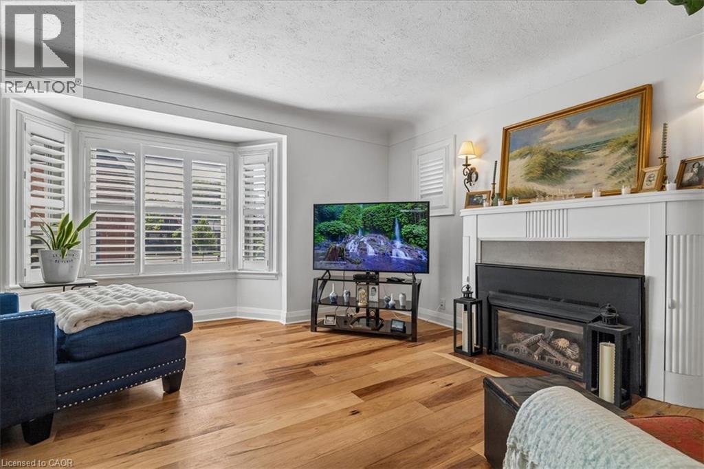 Living area featuring wood-finish flooring, a bay window with white plantation shutters, a decorative fireplace with a mantel, and wall sconces - 573 Beach Boulevard, Hamilton, ON - Indoor Photo Showing Living Room With Fireplace
