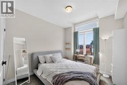 Bright bedroom featuring a gray upholstered bed frame, large windows with operable shutters and natural light, polished concrete flooring, full-length mirror, floor lamp, and city skyline backdrop - 