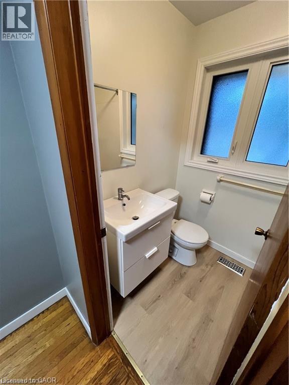 Modern bathroom featuring a white vanity with integrated sink and storage drawer, ceramic toilet, wood-finish flooring, and dual casement windows providing natural light - 683 Coronation Boulevard Unit# 2, Cambridge, ON - Indoor Photo Showing Bathroom