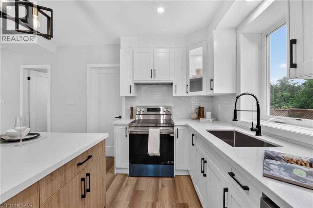 Modern kitchen featuring white shaker cabinetry, stainless steel appliances, a black pull-down faucet, wood-finish flooring, and a large window - 325 Queensdale Avenue E, Hamilton, ON - Indoor Photo Showing Kitchen With Upgraded Kitchen