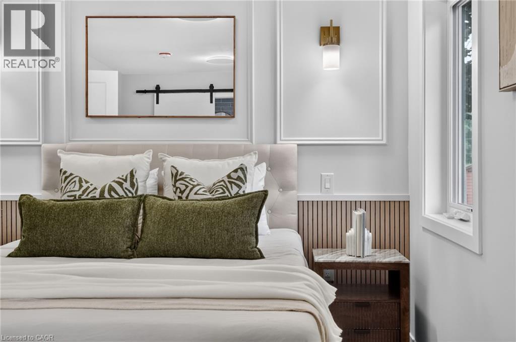 Bedroom featuring wainscoting, an upholstered headboard, a wall-mounted brass light fixture, and a framed mirror - 325 Queensdale Avenue E, Hamilton, ON - Indoor