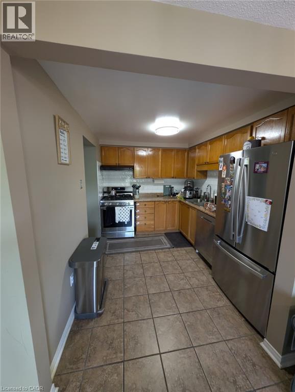 Galley-style kitchen featuring wood-finish cabinetry, stainless steel appliances including a French-door refrigerator, gas range, and tile flooring throughout - 466 Bankside Drive, Kitchener, ON - Indoor Photo Showing Kitchen