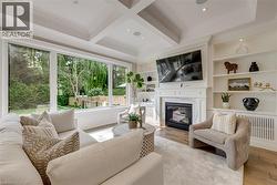 Bright sunroom featuring extensive multi-panel windows framing mature landscaping, coffered ceiling with recessed lighting, wide-plank wood-finish flooring, a gas fireplace with built-in shelving, and - 
