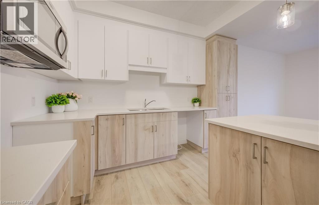 Modern kitchen featuring light wood-finish lower cabinetry and island, white upper cabinetry, white countertops, stainless steel microwave, and wood-finish flooring - 89 Rivergreen Crescent, Cambridge, ON - Indoor Photo Showing Kitchen With Double Sink