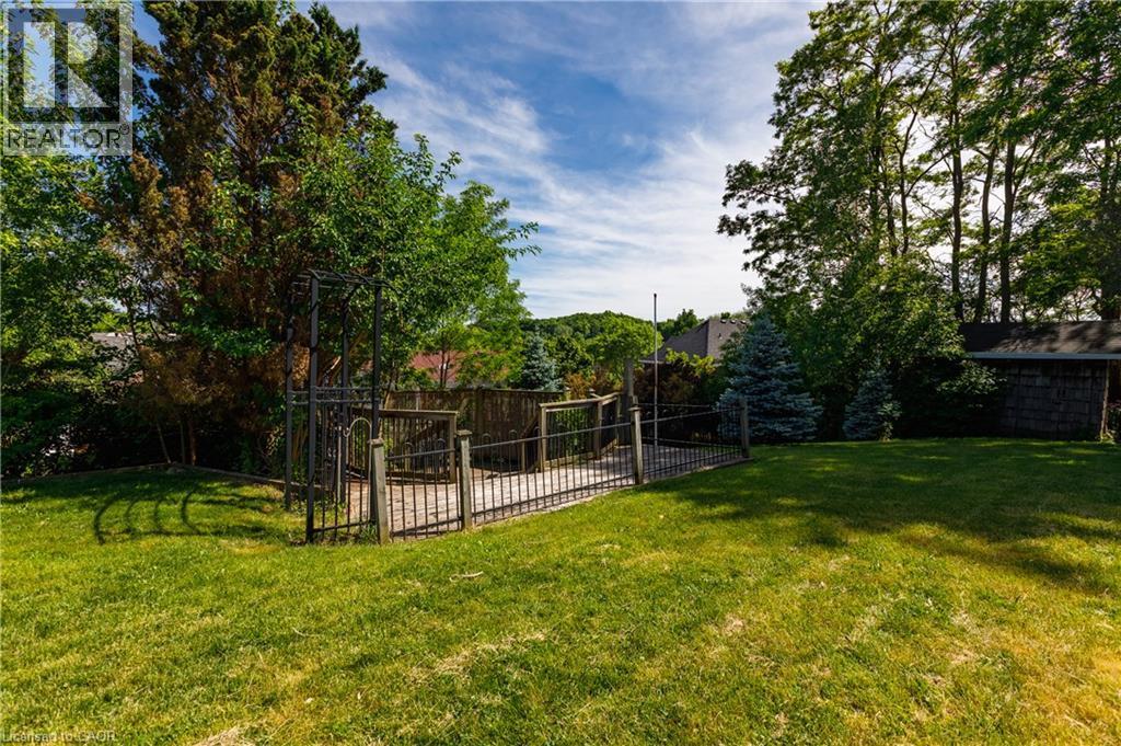 Expansive grassy yard area featuring a fenced deck with a metal gate and surrounding mature trees - 907 Doon Village Road, Kitchener, ON - Outdoor