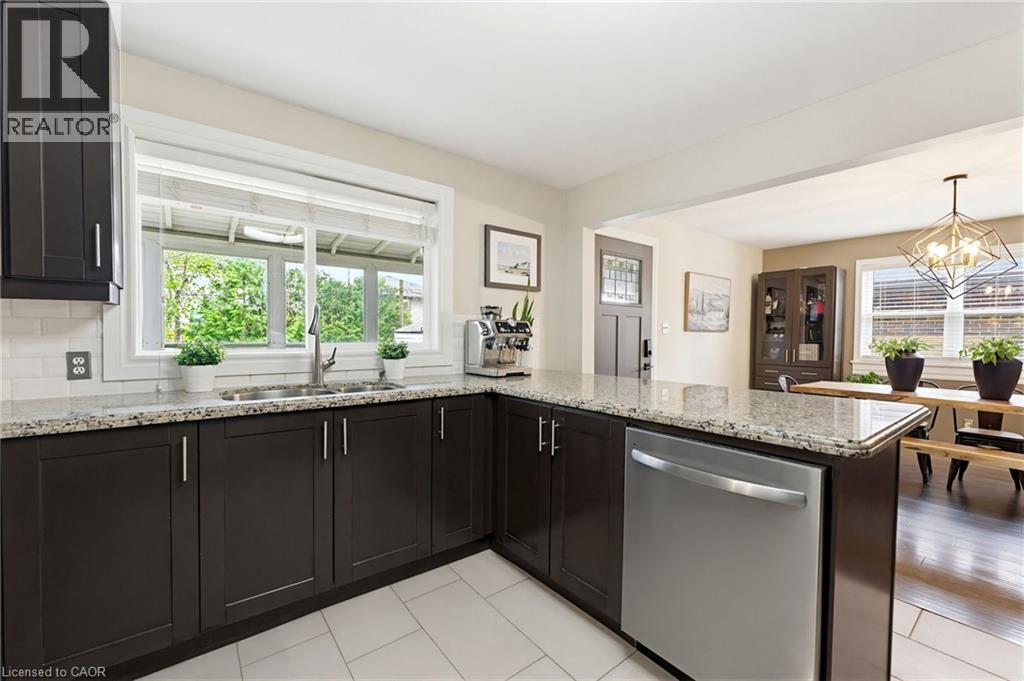 Kitchen featuring dark cabinetry, speckled stone countertops, a stainless steel dishwasher, and a double basin sink beneath a large window - 60 Avenue Road, Cambridge, ON - Indoor Photo Showing Kitchen