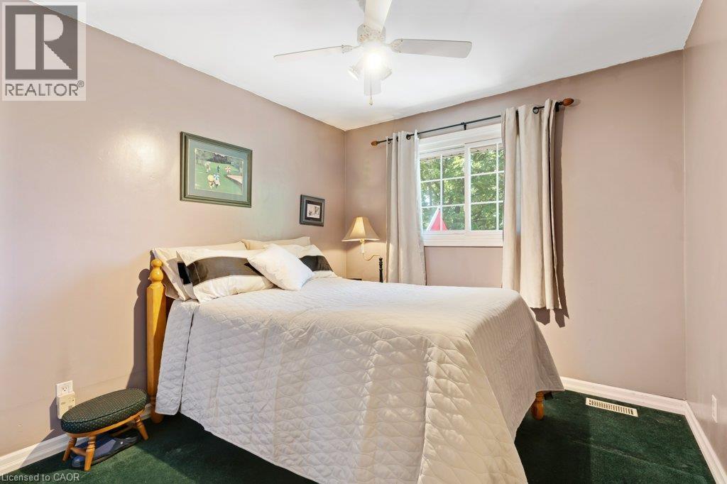 Serene bedroom retreat showcasing soft blush walls, a queen-size bed with quilted bedding, ceiling fan with integrated lighting, large double-hung window with natural light, and dark green-finish floo - 236 Winston Boulevard, Cambridge, ON - Indoor Photo Showing Bedroom