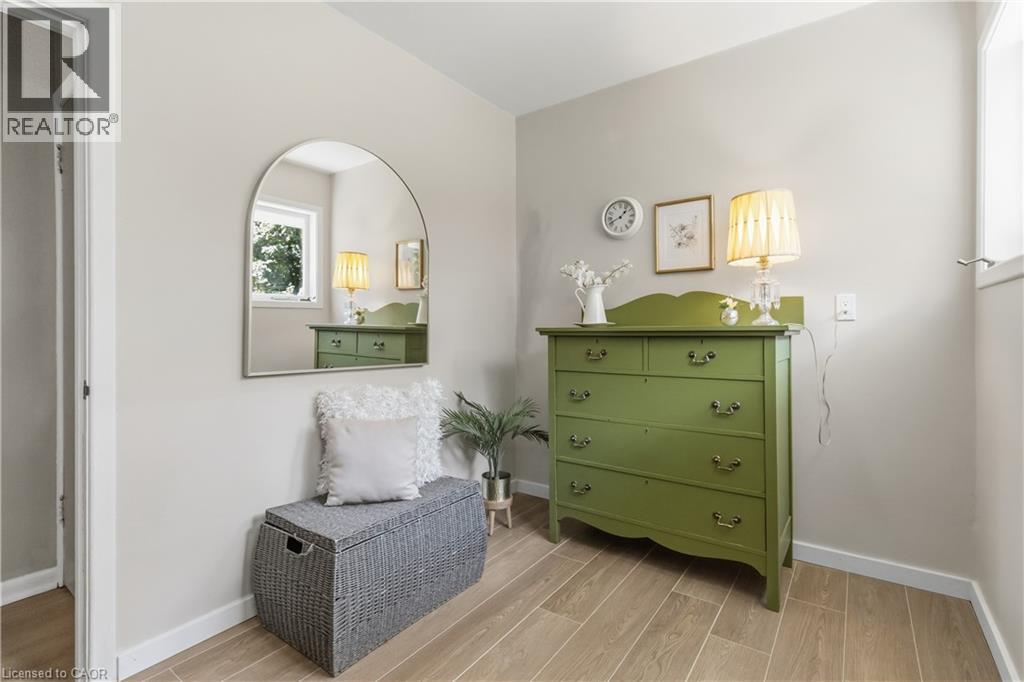 Charming bedroom retreat featuring a sage green dresser with brass hardware, arched mirror with integrated lighting, and warm wood-finish flooring - 974 Unsworth Avenue, Burlington, ON
