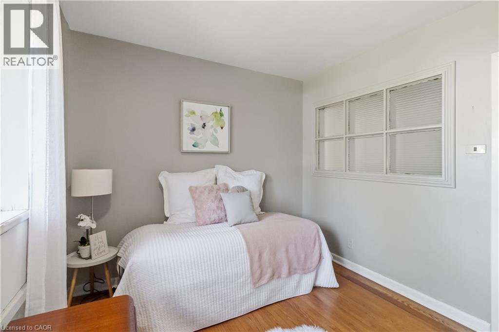 Comfortable bedroom showcasing soft pink and white bedding, a white upholstered headboard, hardwood flooring, gray walls, and a large multi-pane window - 974 Unsworth Avenue, Burlington, ON