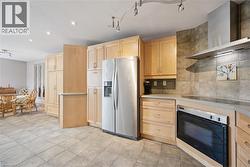 Spacious kitchen featuring natural wood cabinetry, stainless steel refrigerator and built-in wall oven, stone tile backsplash, and tile flooring throughout - 
