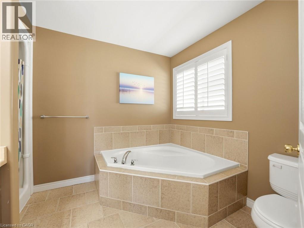 Corner soaking tub with tile surround, white plantation shutters, and light tan wall finishes - 98 Willow Lane, Grimsby, ON - Indoor Photo Showing Bathroom