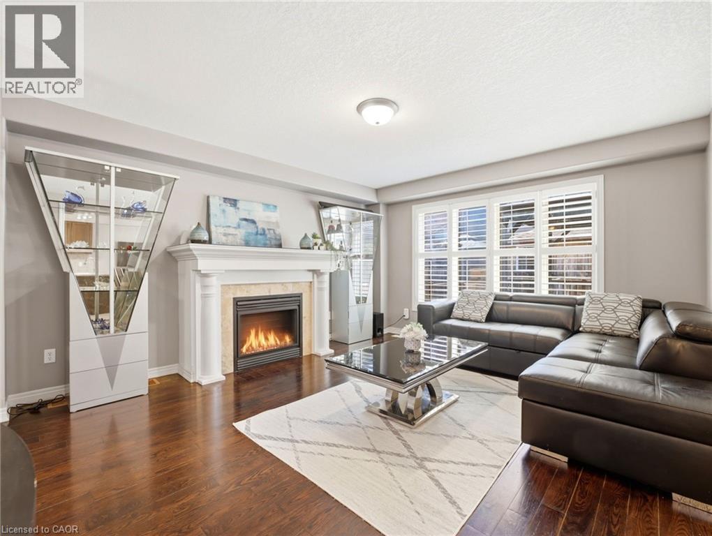 Living area featuring a fireplace with a white mantel and stone surround, wood-finish flooring, a large window with white plantation shutters, and light grey wall paint - 98 Willow Lane, Grimsby, ON - Indoor Photo Showing Living Room With Fireplace