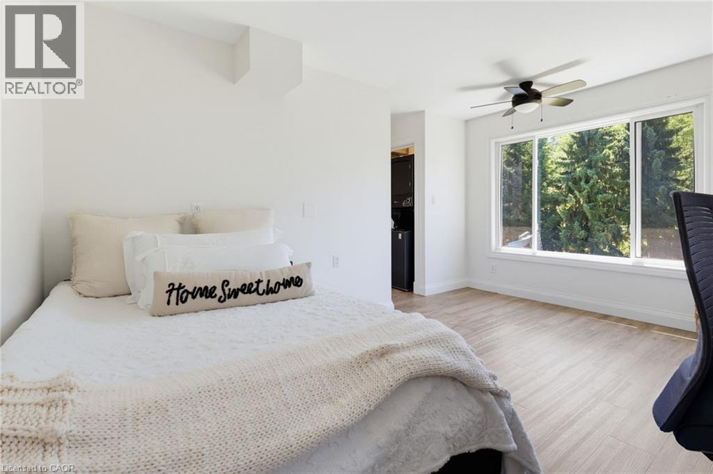 Bright primary bedroom showcasing light wood-finish flooring, large multi-panel windows with natural light, ceiling fan with integrated lighting, and neutral wall finishes - 21 Kerman Avenue, Grimsby, ON