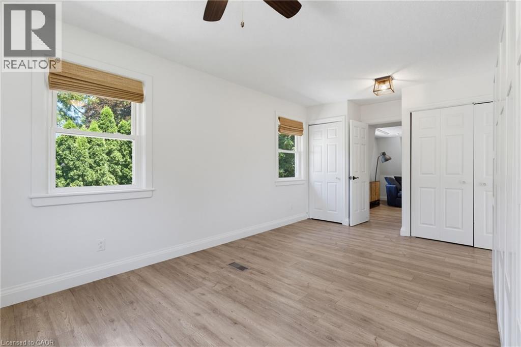 Bright and spacious bedroom featuring hardwood-finish flooring, abundant natural light from dual windows with wood-trimmed frames, white bi-fold closet doors, and a ceiling fan - 21 Kerman Avenue, Grimsby, ON
