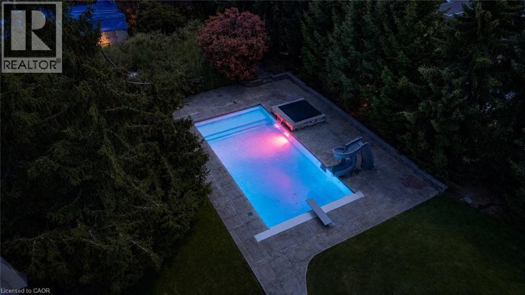 Illuminated resort-style swimming pool with gradient lighting features, complemented by a spacious concrete patio and mature evergreen landscaping - 21 Kerman Avenue, Grimsby, ON