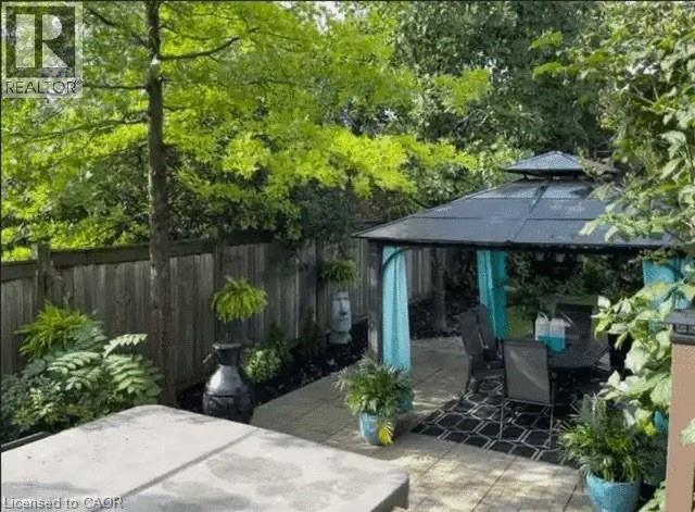 Private patio space featuring a metal-framed gazebo with a dark roof and light blue fabric accents, a paved patio with dark patterned outdoor rug, surrounding lush greenery, and a wooden privacy fence - 4000 Medland Drive, Burlington, ON - Outdoor